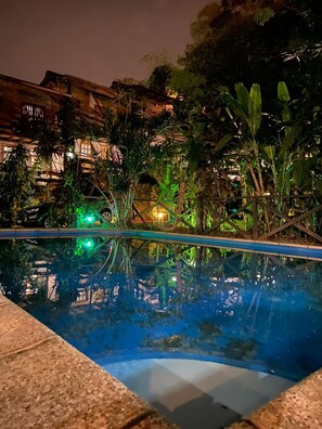 A nighttime pool area with a clear view of the pool's edge, surrounded by lush greenery and a building with warm lighting in the background. - jasy hotel (Puerto Iguazú)