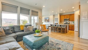 A spacious living room with a large sectional sofa, a blue ottoman, and a view of the kitchen in the background. - Hike, Bike & Swim: Utah Family Home w/ Mtn Views! (Washington)