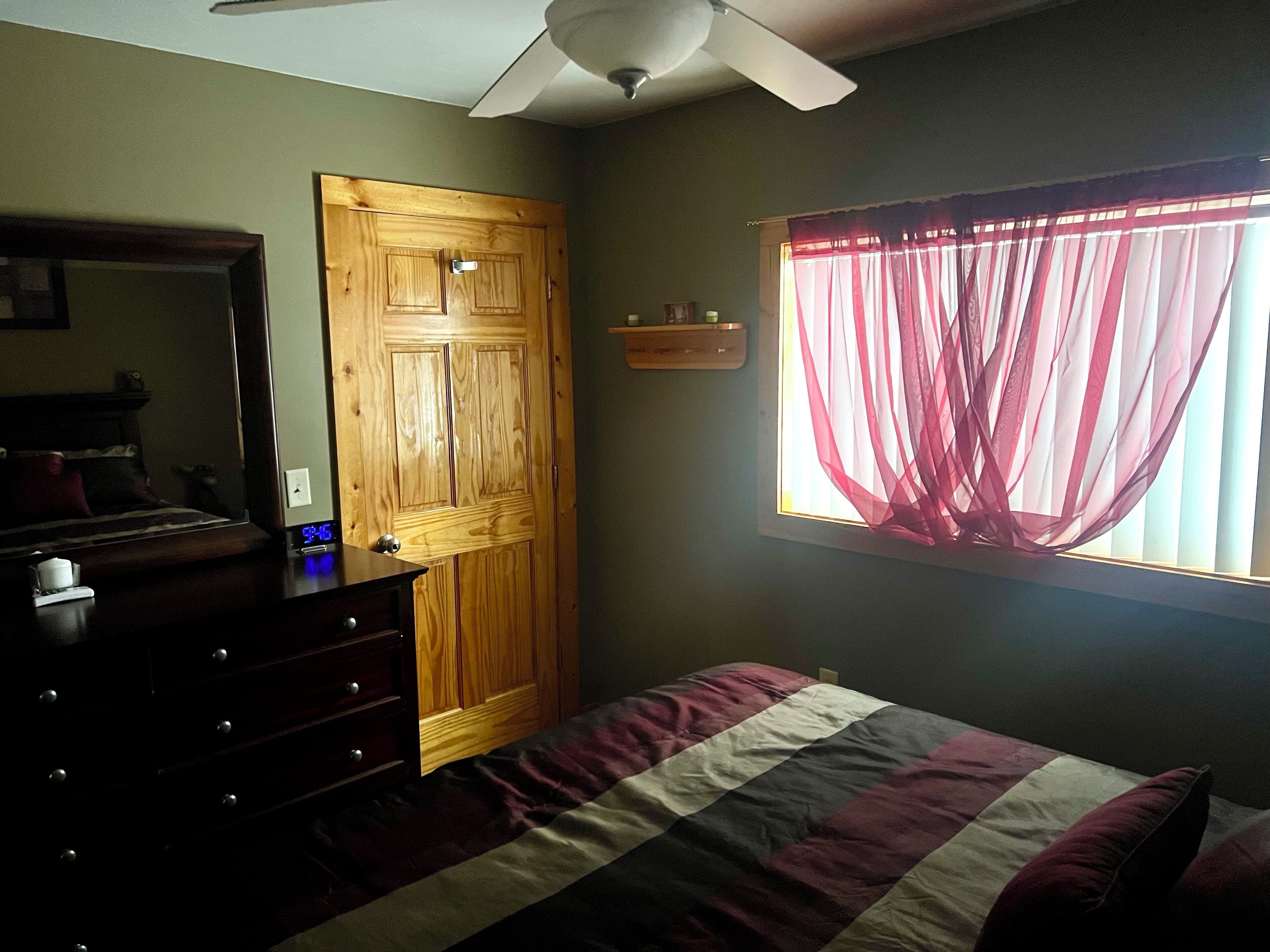 Een slaapkamer met een houten garderobe, een bed met een gestreept dekbedovertrek, een plafondventilator en een raam met dunne gordijnen.