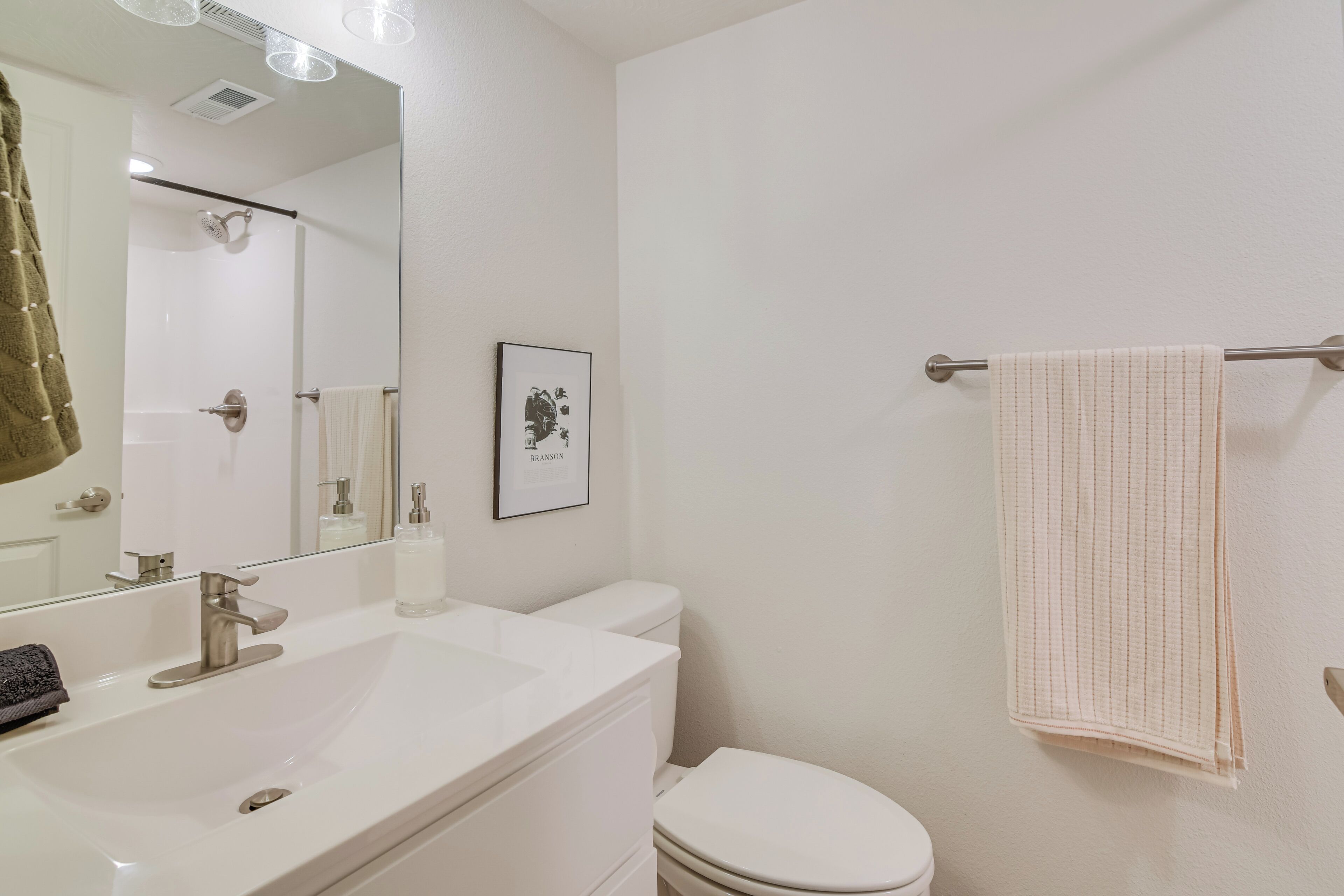 A bathroom with a white sink, mirror, and toilet. There is a towel rack with a towel hanging on it. A framed picture is on the wall.
