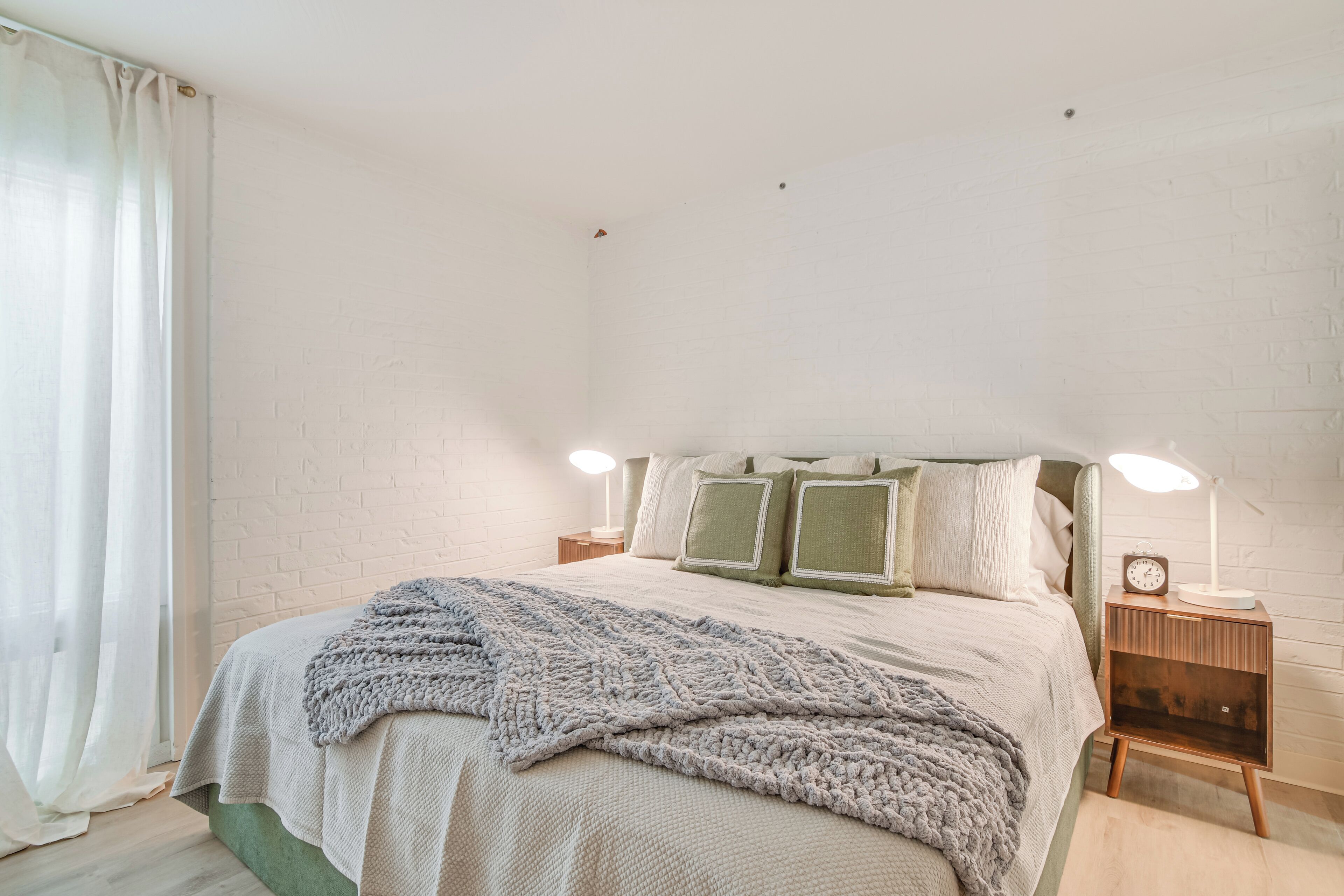 A neatly made bed with a grey blanket, a bedside lamp, and a clock on a wooden nightstand.