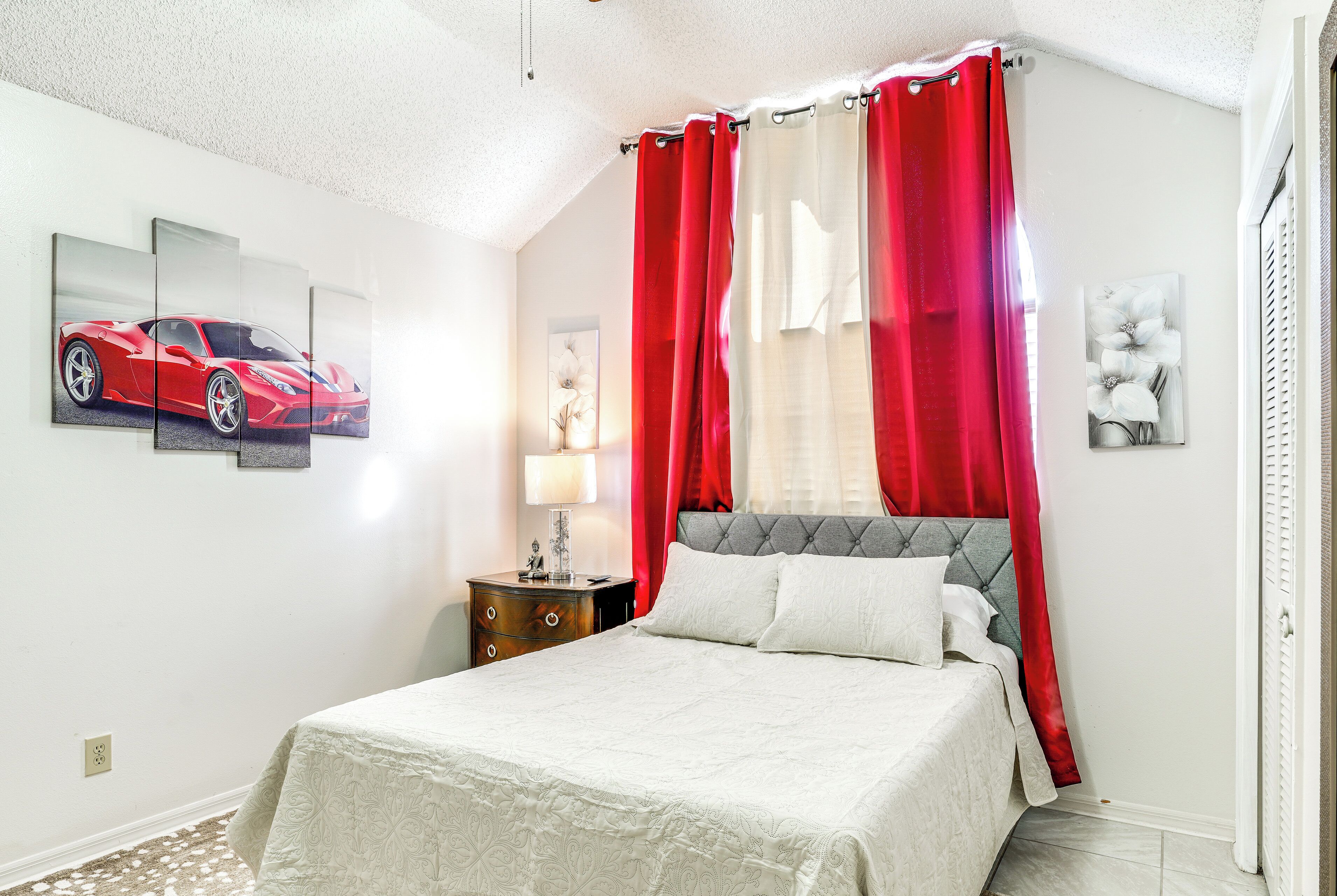 A bedroom with a bed, a nightstand, a lamp, a red and white curtain, and a framed picture of a car.