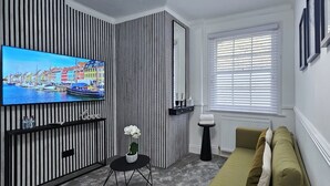 A modern living room with a flat-screen TV, striped wall paneling, a small round table with a potted plant, a sofa, and a window with blinds.