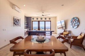 A spacious dining area with a large wooden table, wicker chairs, and a view of the ocean through large windows.