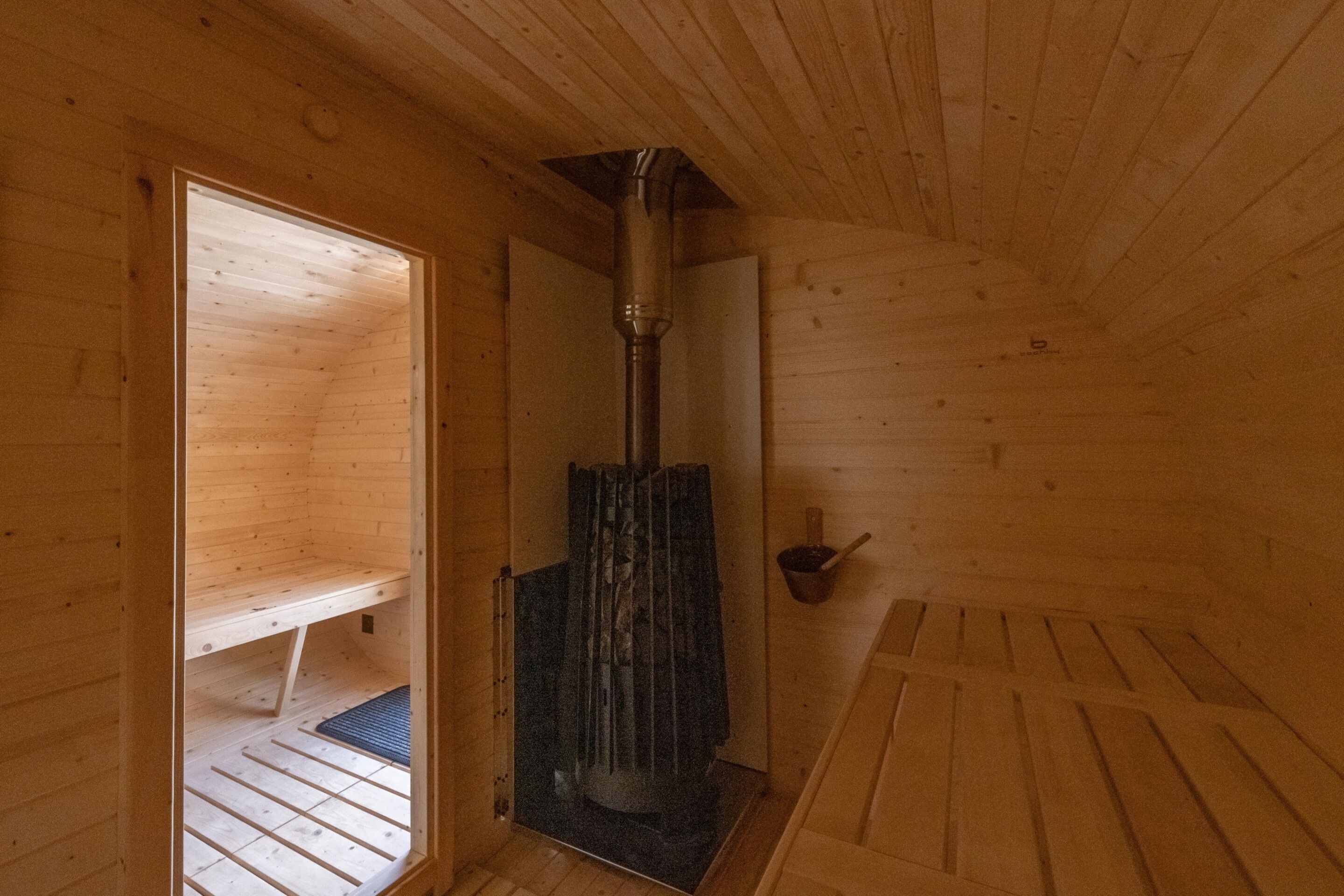 A wooden sauna room with a central heating column, a bench, and a small shelf.