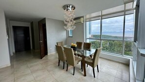 A spacious dining area with a modern chandelier, a large window offering a view of the city, and a door leading to another room. - River Wood I Sunny 1 BDR in Costa del Este (Panama City)