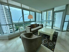 Uma sala de estar moderna com sofá, poltrona e mesa de centro, com grandes janelas que oferecem vista da cidade.