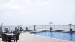 A rooftop pool area with lounge chairs and tables, a clear view of the ocean, and a glass railing. - River Wood I Sunny 1 BDR in Costa del Este (Panama City)
