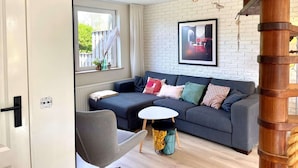 A modern living room with a grey sofa, a small round table, and a chair. There is a window with curtains, a wooden beam structure, and a framed picture on the wall. - Relaxing Holiday Home Near Beach (Sint Maartensvlotbrug)