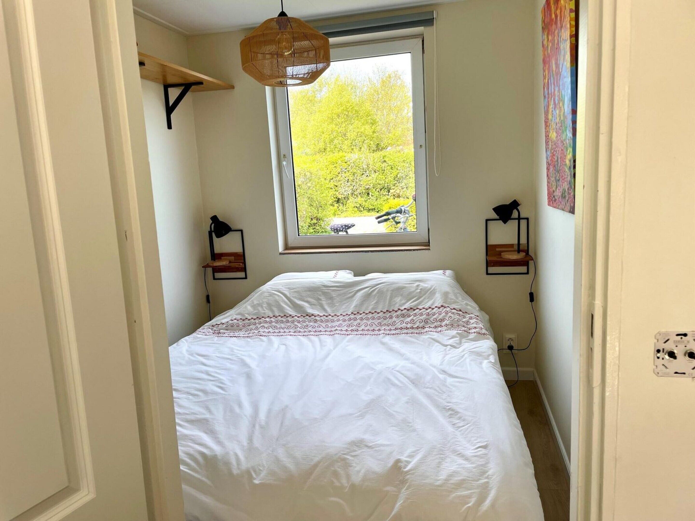 A bedroom with a bed, a window with a view of greenery, a hanging light fixture, and two wall-mounted lamps.