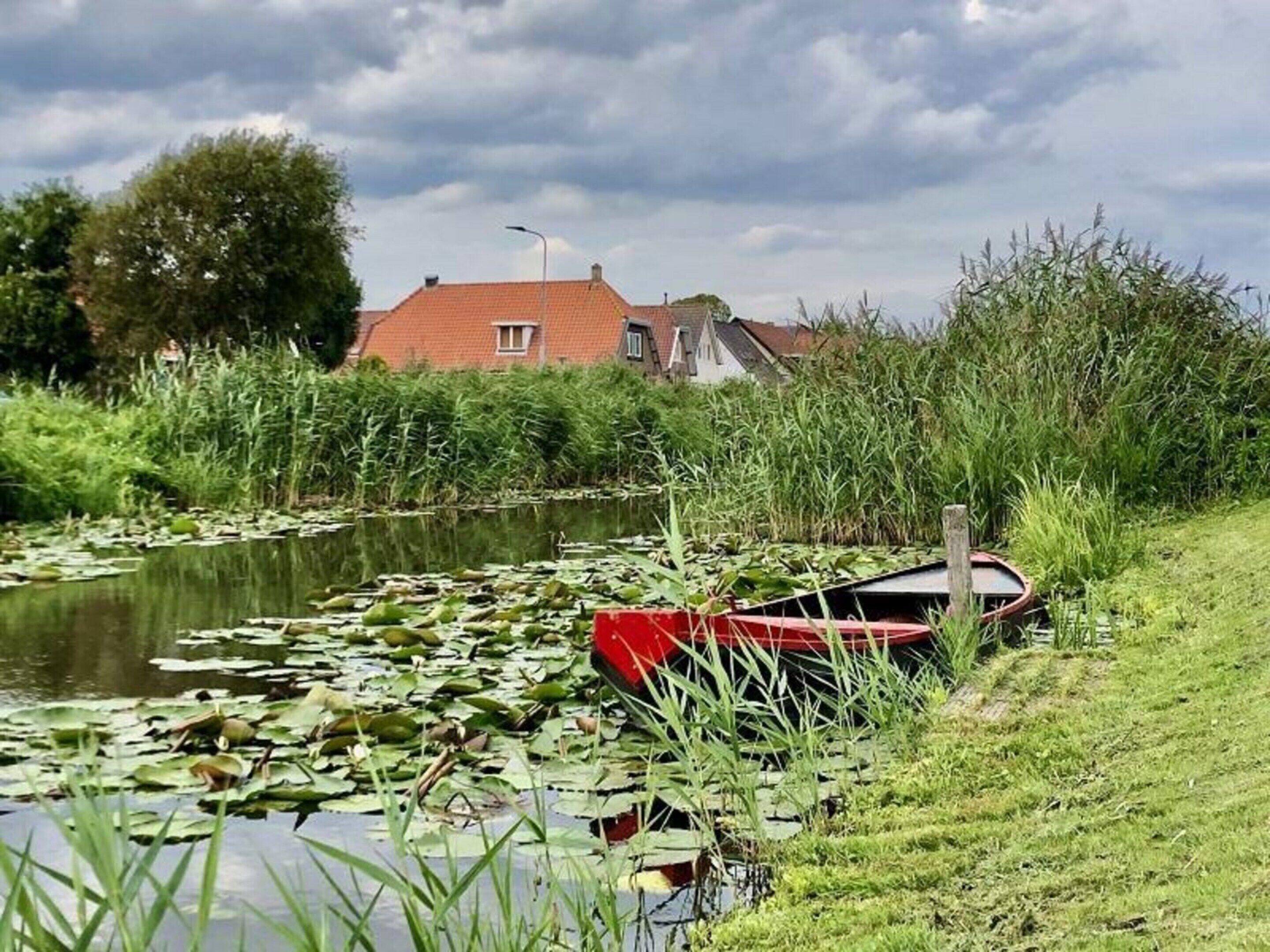 A calm pond with lily pads and a red boat on the grass beside it.