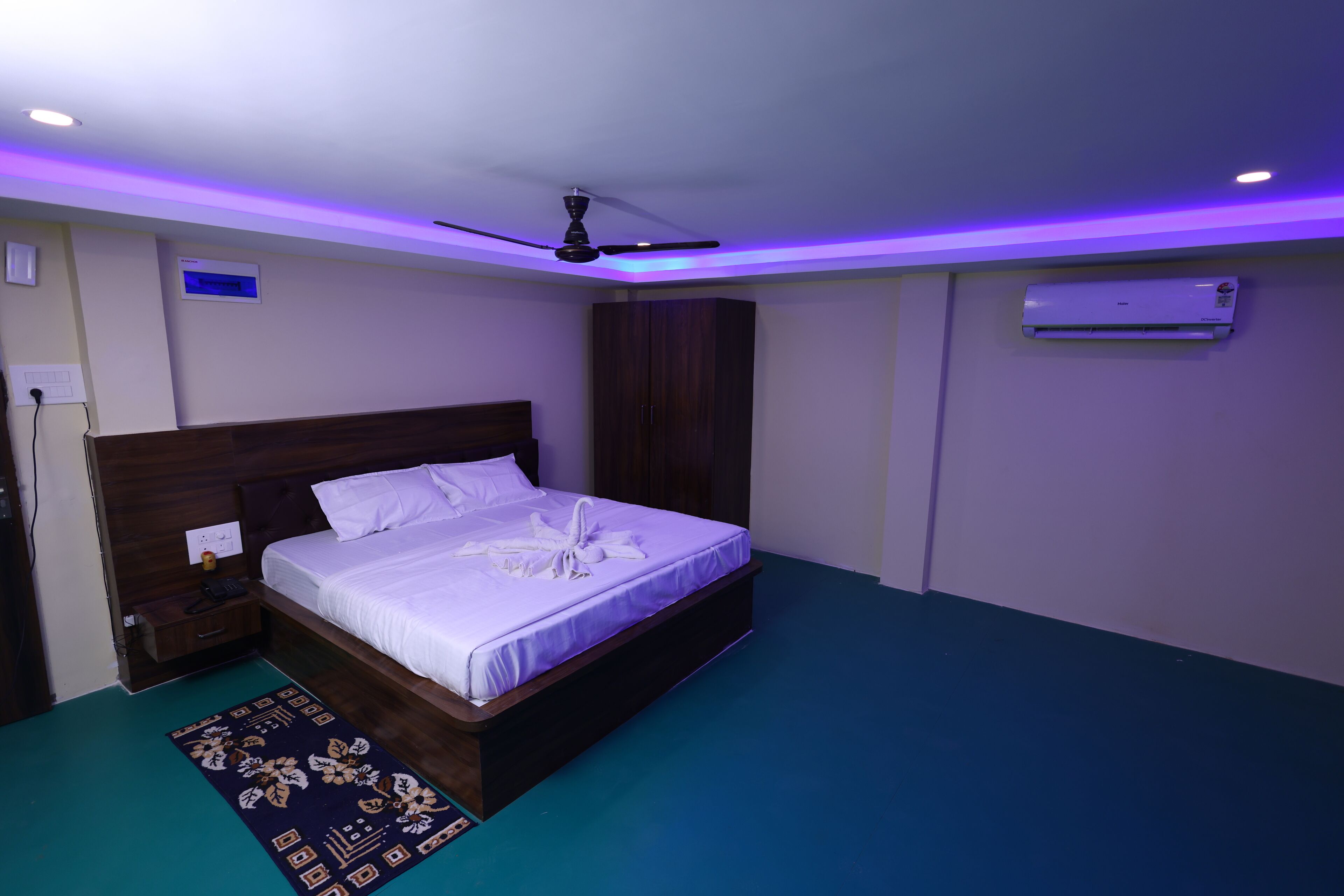 Un lit bien fait, avec une couette blanche et une serviette décorative joliment disposée, une tête de lit en bois, une table de chevet avec une lampe et un ventilateur de plafond.
