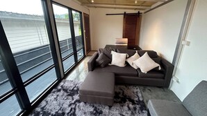 A modern living room with a sectional sofa, a small table, and a large window offering a view of the outdoors. - 1BR Mountain View #2 - 1000mbps WiFi (Ko Pha-ngan)