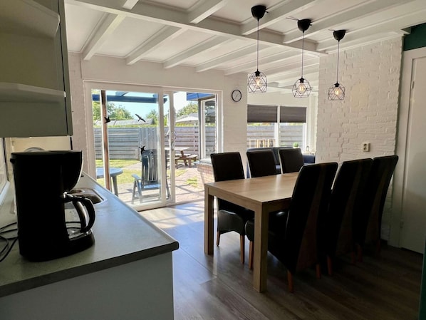 Une cuisine moderne équipée d’une machine à café, d’une table à manger avec des chaises et offrant une vue sur le jardin à travers une porte vitrée.