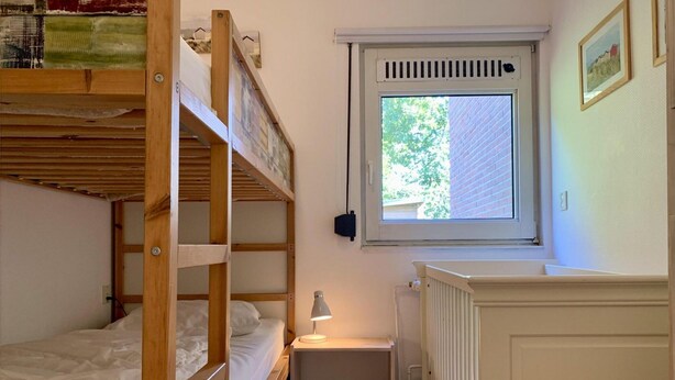 A small, compact room with bunk beds, a window with a view of greenery, a radiator, and a small table with a lamp. - Sunny Garden Retreat Bungalow (Sint Maartensvlotbrug)