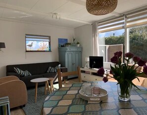 A living room with a sofa, a dining table with a vase of flowers, a cabinet, and a television. - Sunny Garden Retreat Bungalow (Sint Maartensvlotbrug)