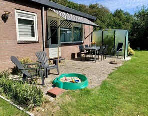 A patio area with a table and chairs, a green play area, and a brick building with a window and door. - Sunny Garden Retreat Bungalow (Sint Maartensvlotbrug)