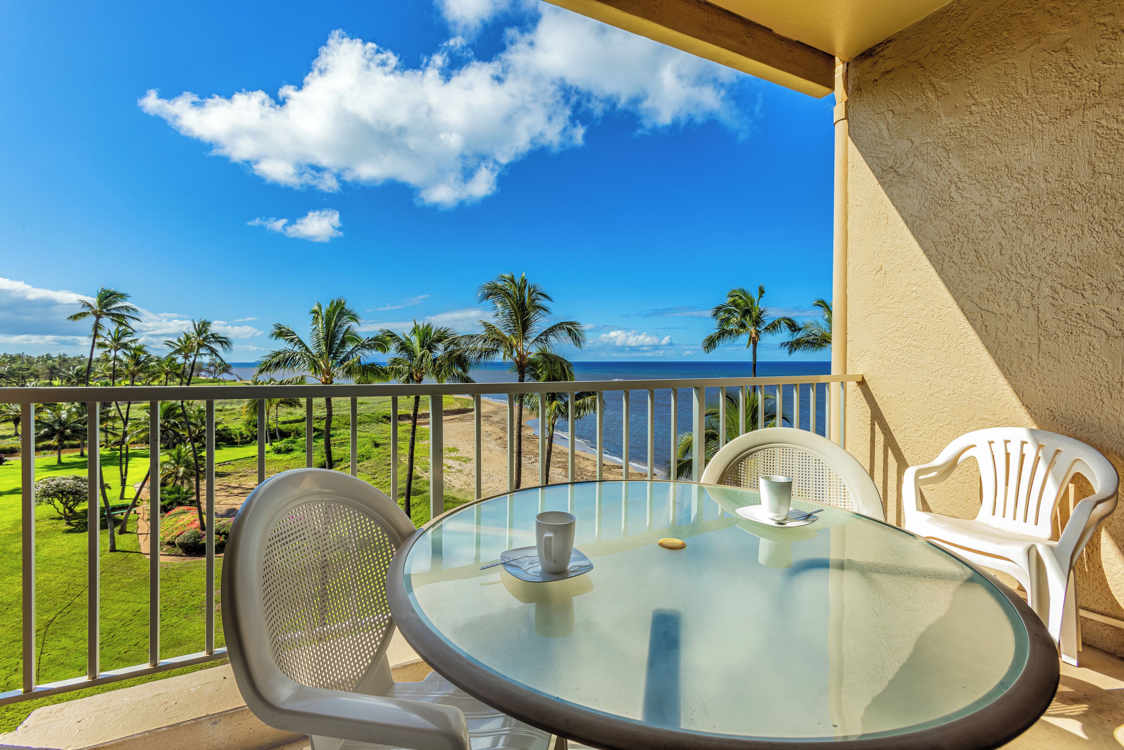 A balcony with a glass table, two white chairs, and a view of a beach and palm trees.
