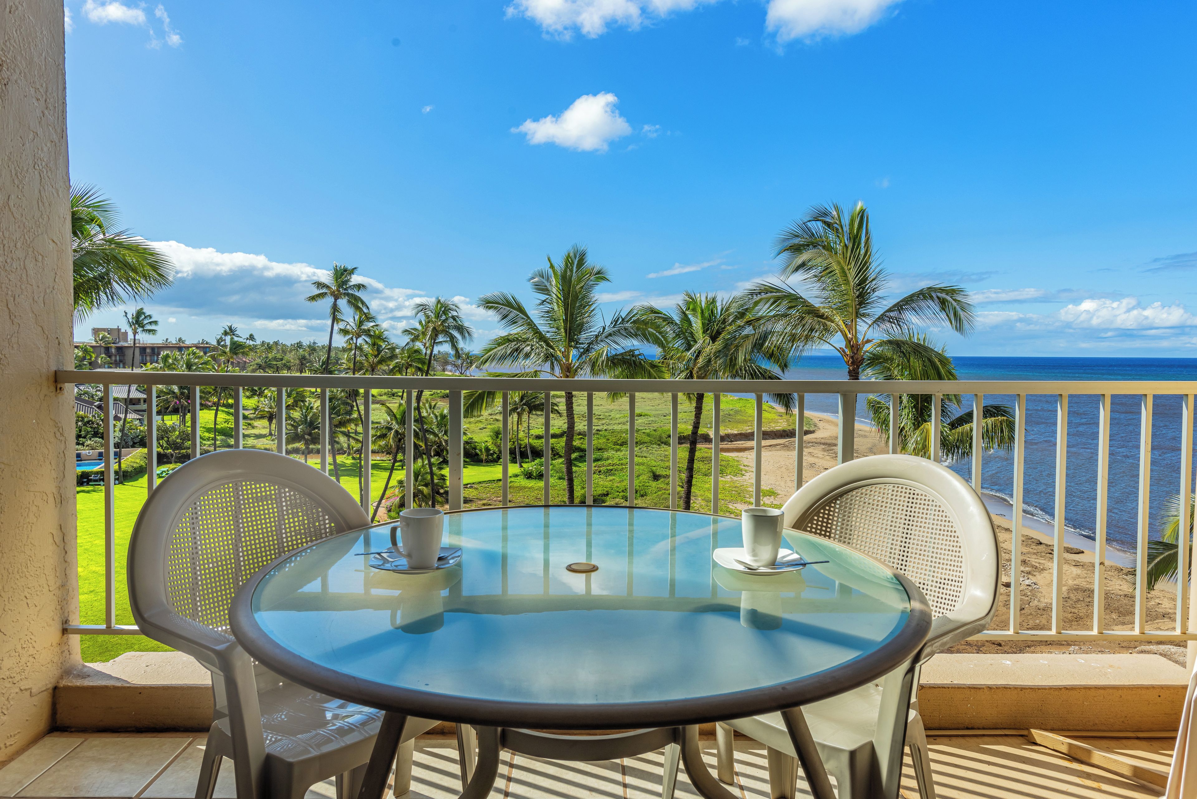 A glass table with two chairs on a balcony overlooking a beach with palm trees and a clear blue sky.