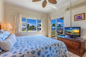Een slaapkamer met een bed, een televisie en een balkon met uitzicht op palmbomen en de oceaan.