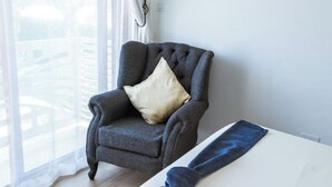 Un fauteuil gris avec un coussin beige, dans une pièce où se trouve une table blanche et d’où l’on aperçoit un bâtiment à travers une fenêtre ornée de rideaux légers.