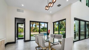 Un espace repas moderne avec une table en verre et quatre chaises, de grandes fenêtres donnant sur une piscine et un jardin, et un luminaire au plafond.