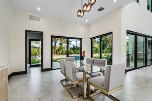 Un espace repas moderne avec une table en verre et quatre chaises, de grandes fenêtres donnant sur une piscine et un jardin, et un luminaire au plafond.