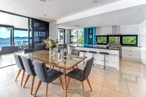 A modern kitchen with a large island, a dining area with a glass table, and a view of the sea. - Edge Apartment 17, Edge Apartment 17 on Hamilton Island (Hamilton Island)