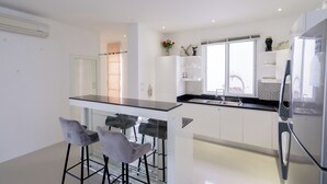 A modern kitchen with white cabinets, a black countertop, and a stainless steel refrigerator. There are two bar stools and a window providing natural light.