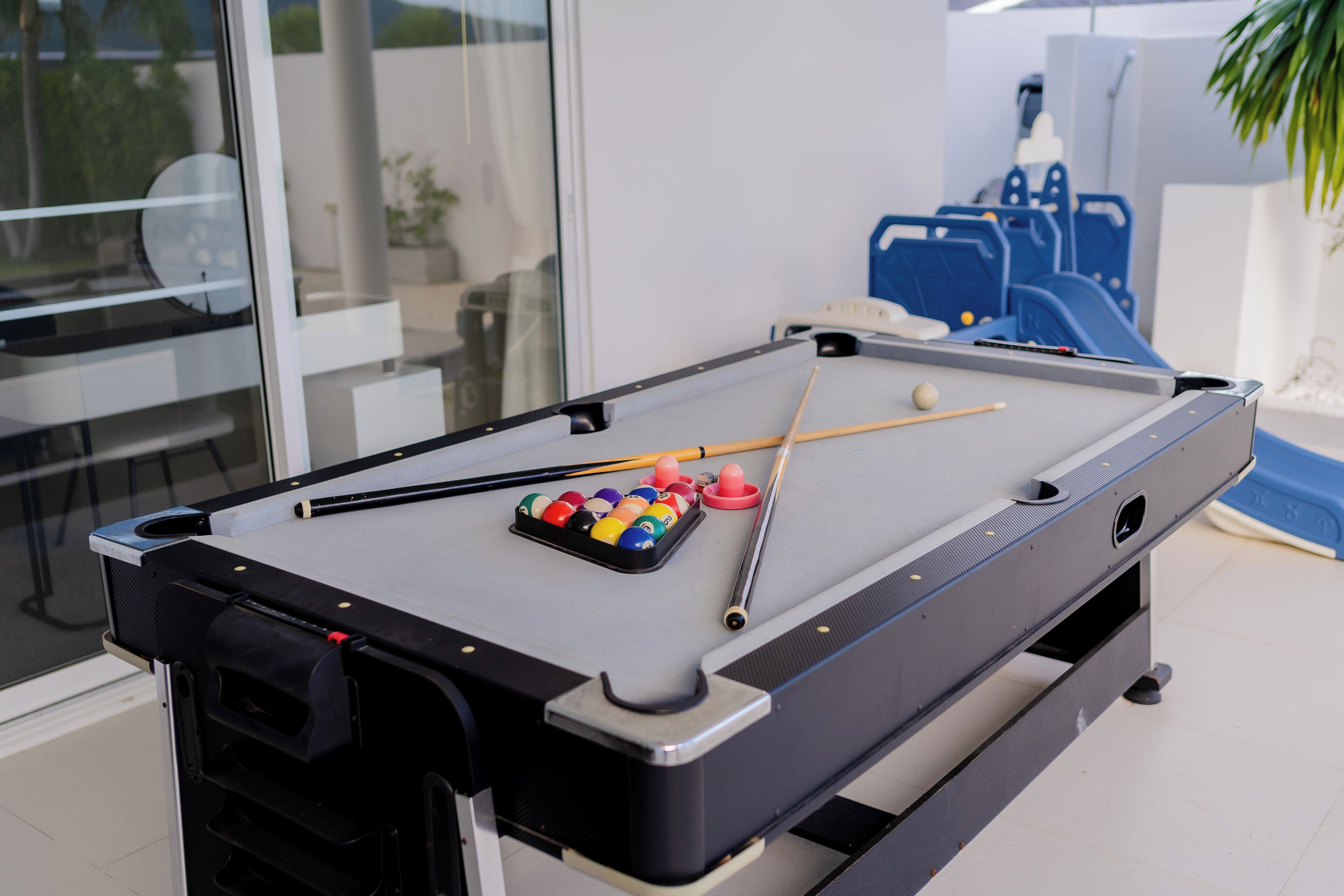 A pool table with balls and cues, a glass partition, and blue chairs in the background.