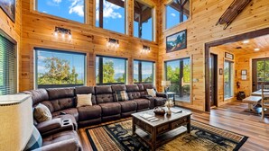A spacious living room with a large brown leather sofa, a coffee table, and a rug. There are multiple windows allowing natural light and views of the outdoors. - Private Indoor Pool • Luxury Lodge • Theater, Firepit (Sevierville)