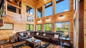 A spacious living room with a large sectional sofa, wooden flooring, and large windows offering views of the outdoors. - Private Indoor Pool • Luxury Lodge • Theater, Firepit (Sevierville)