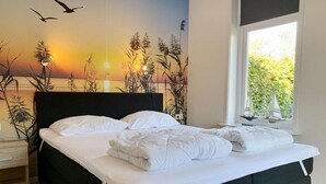 A bedroom with a bed, a nightstand, a window with a view of greenery, and a wall mural of a sunset. - Great Bungalow Near the Beach and sea (Sint Maartensvlotbrug)