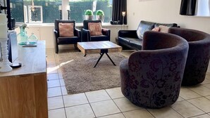 A living room with a sofa, armchairs, a coffee table, and a TV. There are plants and a painting on the wall. - Great Bungalow Near the Beach and sea (Sint Maartensvlotbrug)
