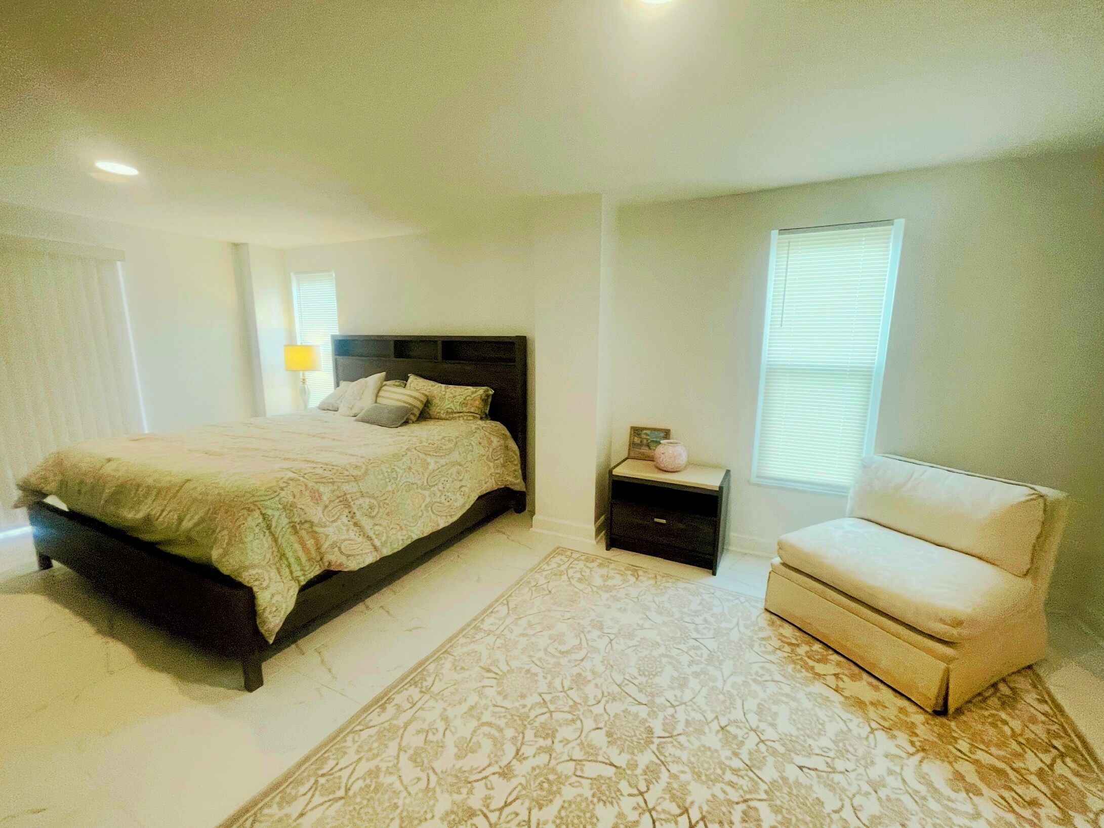 A bedroom with a large bed, a nightstand, a chair, and a window with blinds.
