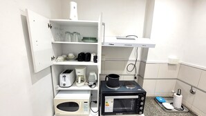A kitchenette with a microwave, toaster, coffee maker, and a small oven. There is a shelf with cups, glasses, and a kettle. A power strip is visible on the wall. - Luminoso, muy Completo y Bien Ubicado (Buenos Aires)