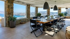 A dining area with a long table, black chairs, and hanging pendant lights. The room has stone pillars and large windows with a view of the sea and hills. - Villa Tavani Agios Ioannis Mykonos (Mykonos)