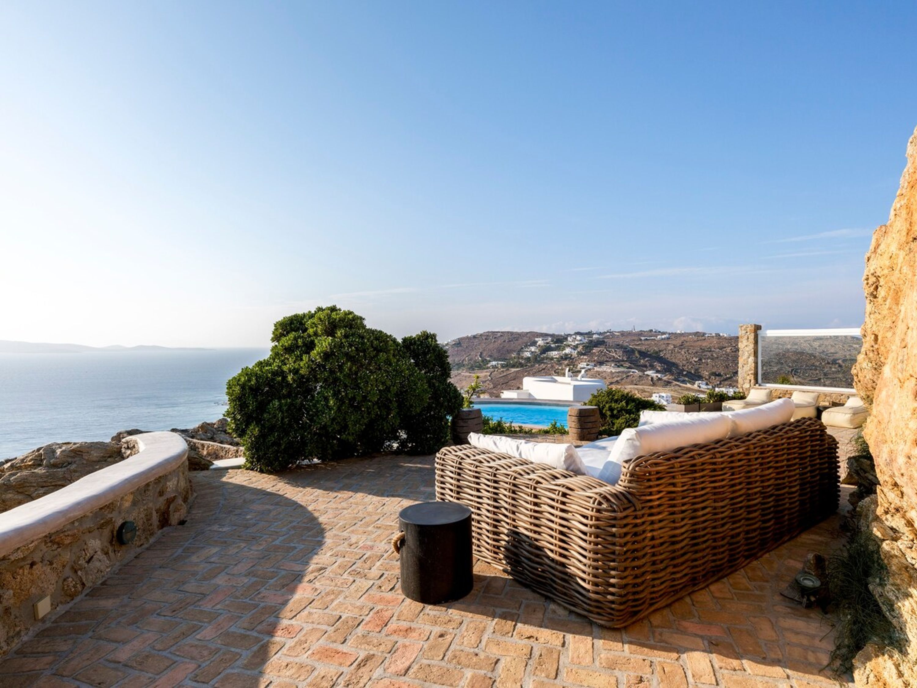 A stone patio with a wicker sofa and a small table, overlooking a coastal landscape with a clear view of the sea and distant hills.