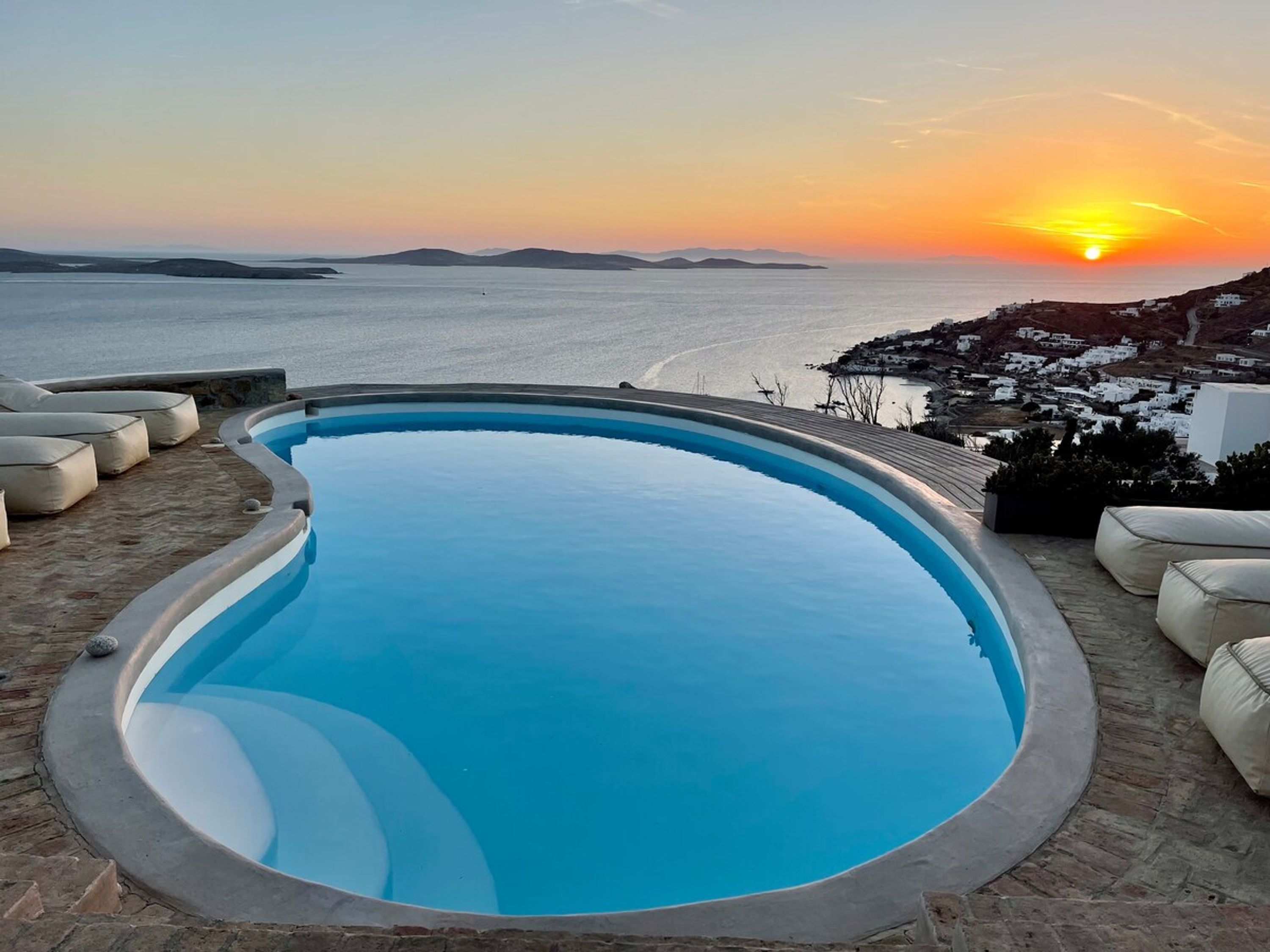 A swimming pool with a curved edge, surrounded by lounge chairs and a view of the sea and distant hills during sunset.