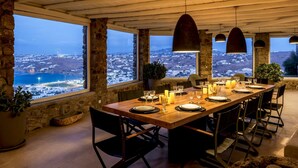 A dining area with a long table set for six, large windows offering a view of a coastal landscape, stone walls, and hanging pendant lights. - Villa Tavani Agios Ioannis Mykonos (Mykonos)