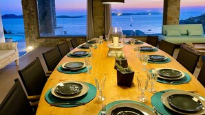 Een eetruimte met een houten tafel gedekt voor een maaltijd, grote ramen met uitzicht op zee en verre eilanden, en een stenen pilaar op de achtergrond.