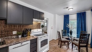 A kitchen with dark cabinets, a white stove, and a dining area with a glass table and blue curtains. - Pet-friendly Furnished Apt Near Emu Free Parking 2 Bedroom Apts (Ypsilanti)