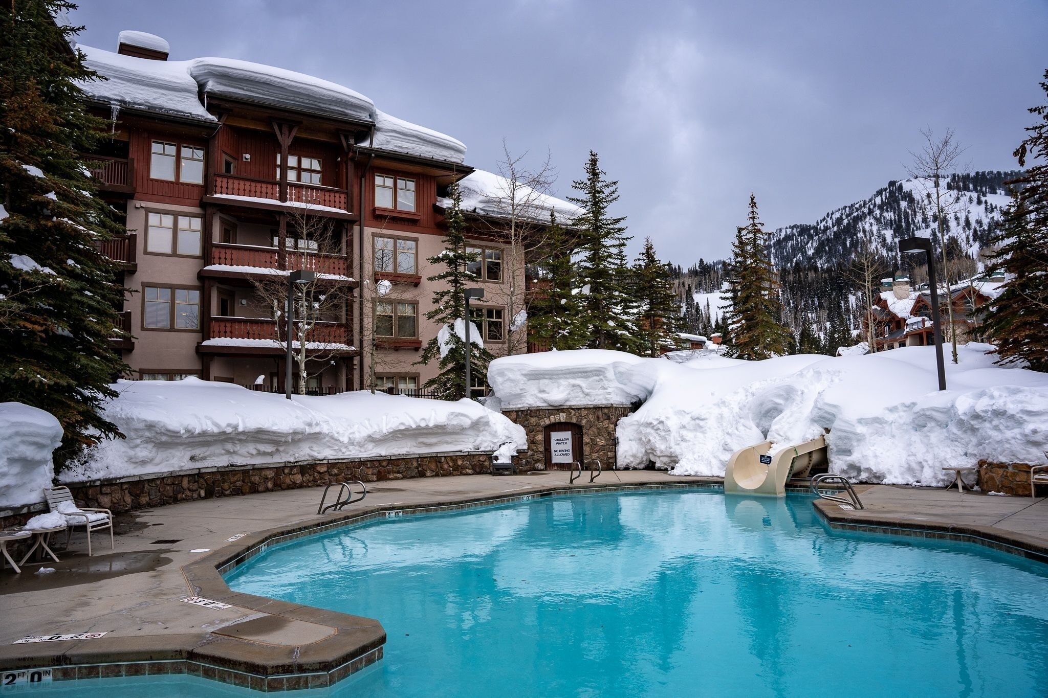 Un área de piscina con una piscina azul cristalina, rodeada de edificios y árboles cubiertos de nieve.