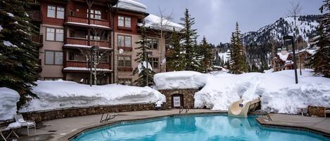 Un área de piscina con una piscina azul cristalina, rodeada de edificios y árboles cubiertos de nieve.
