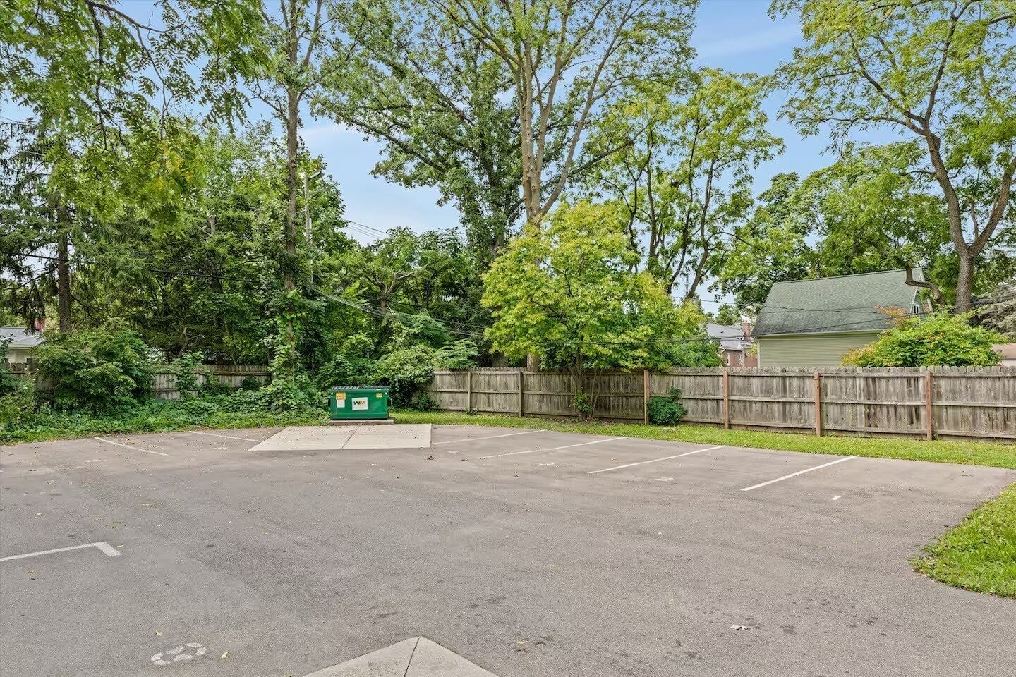 A parking lot with a green dumpster, surrounded by trees and a wooden fence.