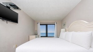 A hotel room with a bed, a flat-screen TV, a nightstand, a lamp, a window with a view of the sea, and a ceiling fan. - Island Sunrise 663 2 Bedroom Condo (Gulf Shores)