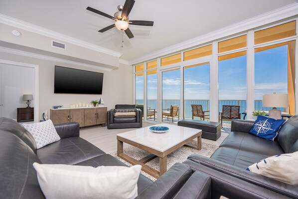 A spacious living room with a flat-screen TV, leather sofas, a coffee table, and a ceiling fan. Large windows offer a view of the ocean. - Phoenix Gulf Towers 1007 3 Bedroom Condo (Orange Beach)