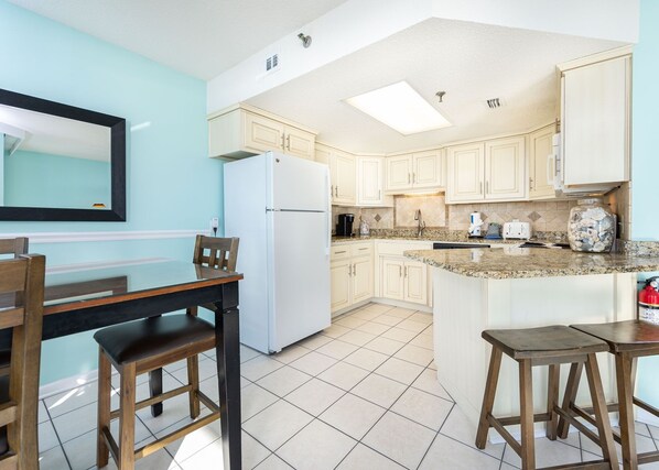 Una cocina con armarios de color claro, un refrigerador blanco, una encimera de granito y una zona de comedor con una mesa y sillas.