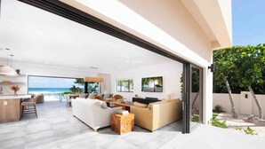 A spacious living area with a large window overlooking the ocean, a dining area, and a kitchen. - Halcyon Point: New Luxurious Oceanfront Villa w/ Pool, Spa + Tanning Terrace (Grand Cayman)