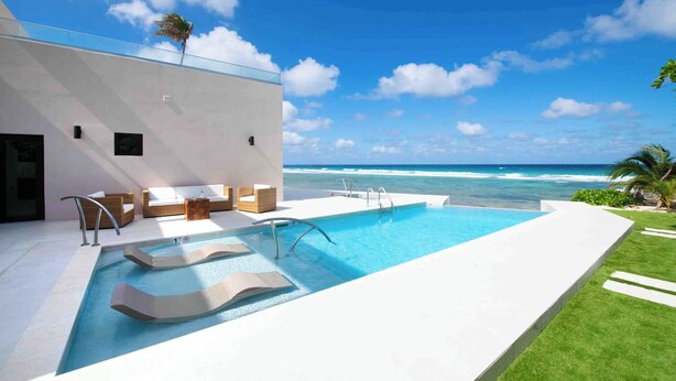 A modern pool area with a view of the ocean, a white building, and outdoor seating. - Halcyon Point: New Luxurious Oceanfront Villa w/ Pool, Spa + Tanning Terrace (Grand Cayman)